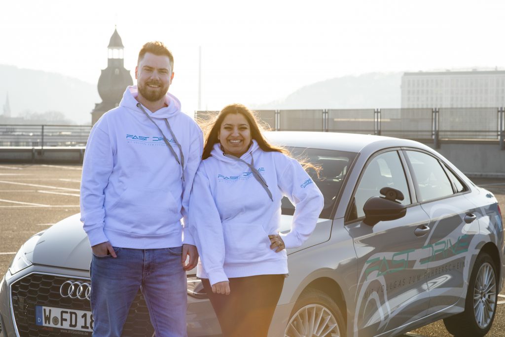 Zu sehen sind die beiden Besitzer von Fast Drive Wuppertal, einer Fahrschule in Wuppertal Elberfeld. Gemeinsam stehen Sie lächelnd vor dem Fahrschulauto auf einem Parkplatz.